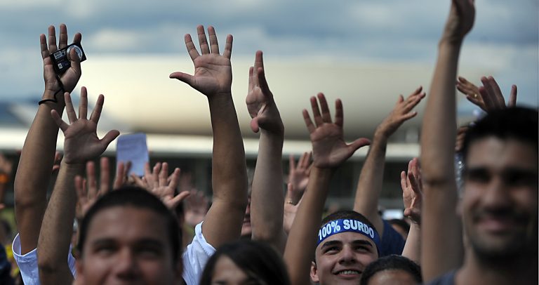 Foto de várias mãos levantadas para o alto. No meio, há um menino com uma faixa amarrada na cabeça escrito "100% surdo"