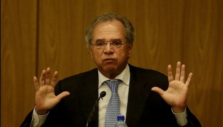 Foto do ministro da Economia, Paulo Guedes, que tem cabelos lisos e grisalhos, usa óculos, veste terno escuro, camisa branca e gravata azul. Está com as duas mãos abertas e erguidas na altura dos ombros.