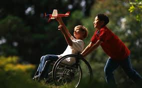 Dois meninos de aproximadamente sete anos de idade estão brincando em um ambiente aberto, com gramado e luz do sol suave. Um deles, na cadeira de rodas, segura bem alto um aviãozinho de brinquedo, e o outro empurra a cadeira de rodas. Os dois estão sorrindo.