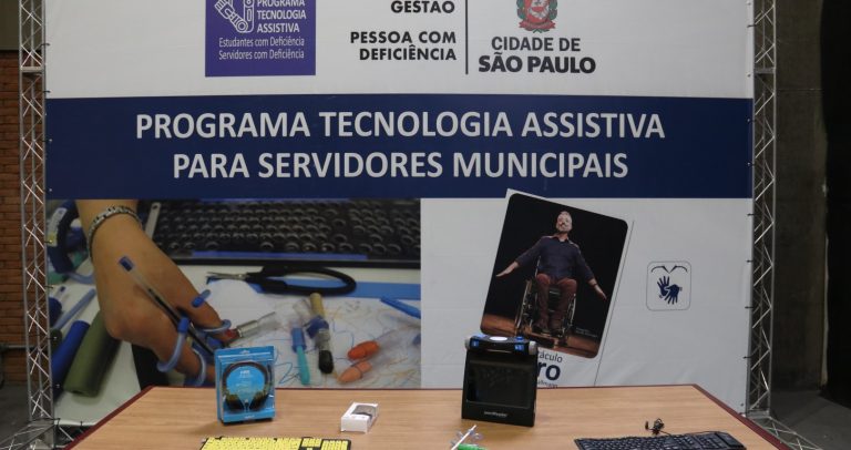 banner ao fundo com informações do Programa Tecnologia Assistiva para Servidores Municipais e da peça Ícaro. Sobre a mesa, alguns equipamentos de tecnologia assistiva.
