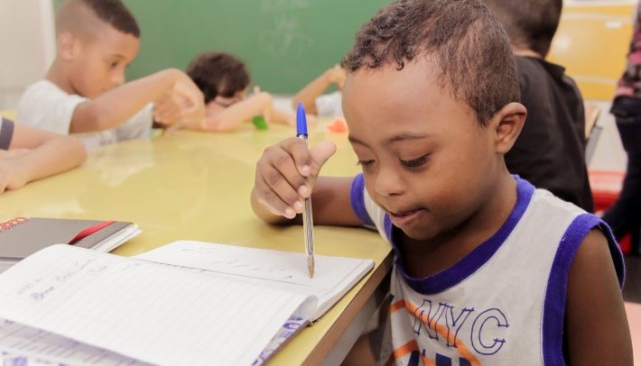 Foto de um menino com Síndrome de Down. Ele está sentado junto com outras crianças em volta de uma grande mesa amarela. Ele é negro, tem cabelos curtos, enrolados e escuros. Com a mão direita, segura uma caneta esferográfica azul e escreve em um caderno. Ele usa um uniforme escolar e olha para o caderno.