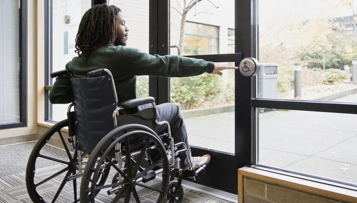 Foto de um homem em uma sala fechada. Ele está sentado em uma cadeira de rodas e ergue a mão direita em direção a um botão exisstente em uma porta à frente dele. O botão contém o símbolo universal da pessoa com deficiência.