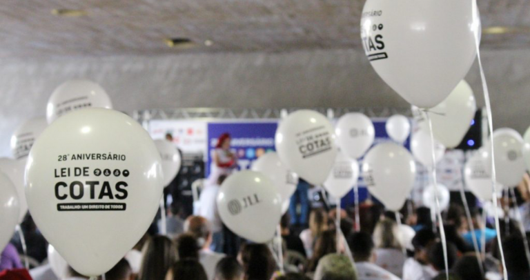 Foto tirada nas comemorações do 28º aniversário da Lei de Cotas, na capital paulista. Sobre uma porção de balões brancos, está escrito em letras pretas: “Lei de Cotas – Aniversário de 28 anos”. Ao fundo, há uma multidão desfocada de pessoas.