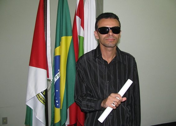 Foto de um homem segurando um diploma de formatura. Ele está de pé, quase ao centro, retratado da cintura para cima. Tem cabelos pretos e curtos, pele branca, usa óculos escuros e camisa preta listrada. Ao fundo, há quatro bandeiras. Uma delas é a bandeira nacional e as outras estão ilegíveis.