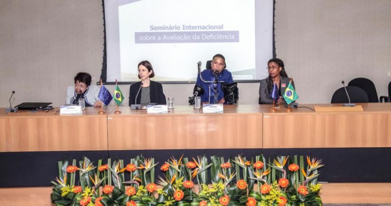 Foto de três mulheres e um homem, cada qual com um microfone. A primeira pessoa da esquerda é Izabel Maior. Ela tem cabelos pretos e curtos e usa óculos. Todos estão sentados em frente a uma mesa. Ao fundo, uma projeção diz: "Seminário Internacional sobre a Avaliação da Deficiência". À frente, há um arranjo de flores.