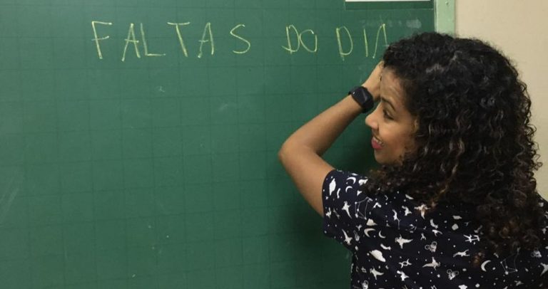 Em uma sala de aula, uma professora escreve em um quadro negro as palavras "faltas do dia". Ela tem cabelos pretos encaracolados e usa uma blusa preta.