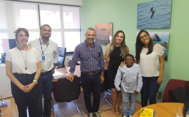 Foto de sete pessoas, homens e mulheres, de pé em uma sala. Uma delas é uma pessoa com nanismo.