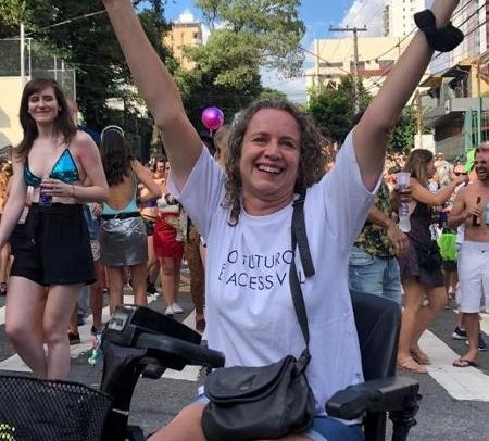 Foto da publicitária Mila Guedes em um bloco de rua. Ela sorri, com os braços erguidos. A moça está sentada em uma cadeira motorizada e usa uma camiseta na qual está escrito "O futuro é acessível".