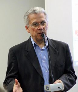 Foto de Heleno Rodrigues Corrêa Filho, da cintura para cima. Ele segura um microfone. Usa terno preto e camisa azul. Ao fundo, há uma parede branca.