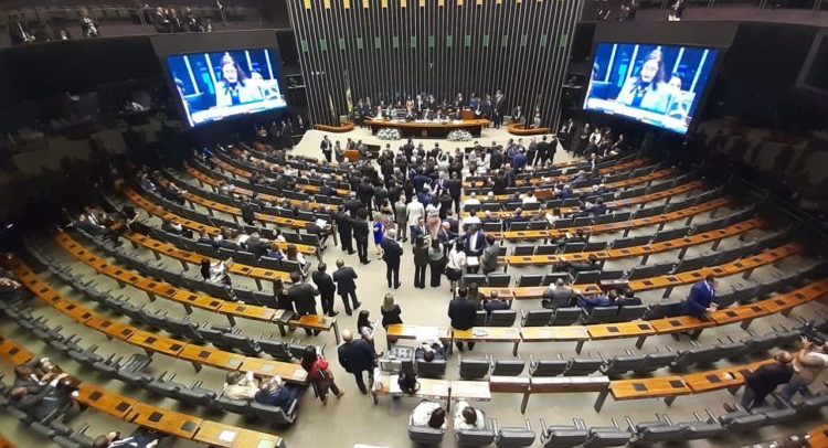 Foto de dentro da plenária. Deputados conversam no corredor, entre as fileiras de mesas.