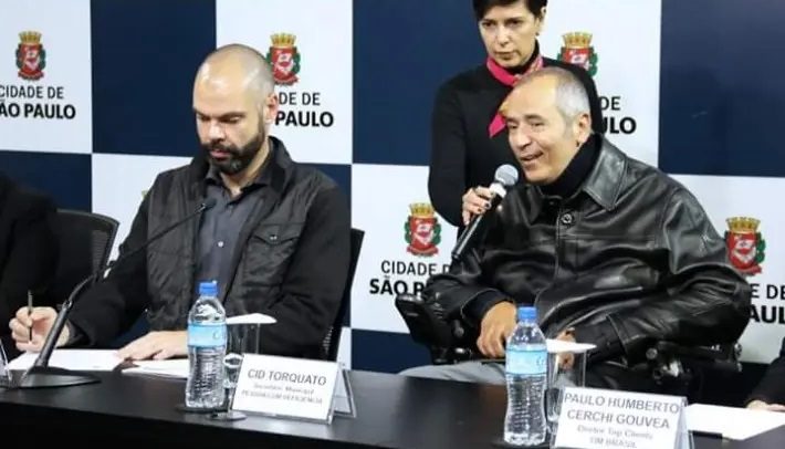O secretário municipal da Pessoa com Deficiência de São Paulo, Cid Torquato, que é tetraplégico, está sentado na cadeira de rodas e fala ao microfone, segurado pela coordenadora de comunicação da SMPED, Ciça Cordeiro, que está em pé. Ao lado deles, está o prefeito Bruno Covas.