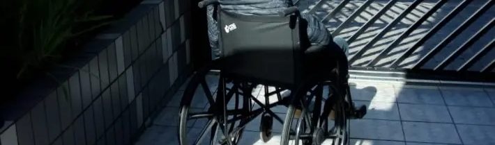 Foto de uma idosa em uma cadeira de rodas. Ela está de costas, na área externa de um edifício, ao lado de um jardim, tomando sol.