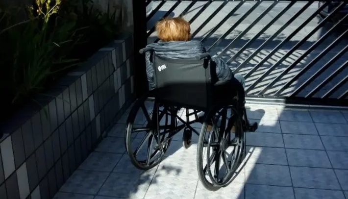 Foto de uma idosa em uma cadeira de rodas. Ela está de costas, na área externa de um edifício, ao lado de um jardim, tomando sol.