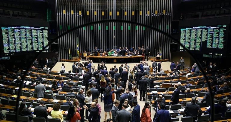 Foto de auditório do Congresso Nacional. Na parte inferior, à esquerda e à direita, estão as mesas dos deputados e senadores. Ainda na parte inferior, ao centro, estão os deputados e senadores em pé conversando no corredor. No canto superior esquerdo e direito, estão as telas que mostram as votações. Na parte central superior, tem a mesa do congresso.