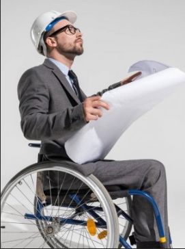 Foto de um homem sentado em uma cadeira de rodas. Ele olha para cima e segura a planta de um edifício. Usa um capacete de proteção, óculos e terno.