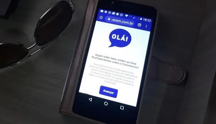 Foto de um smartphone sobre uma mesa, ao lado de um óculos de sol. Na tela do smartphone, a home do site ‘ParaTodosVerem Coronavírus’.