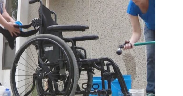 Foto de uma cadeira de rodas. À direita, uma mulher aponta uma mangueira de lavagem para a cadeira.