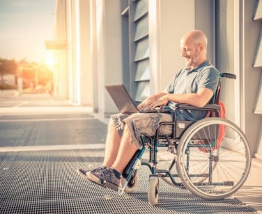 Foto de um cadeirante. Ele está sentado em um ambiente externo e segura um notebook no colo.