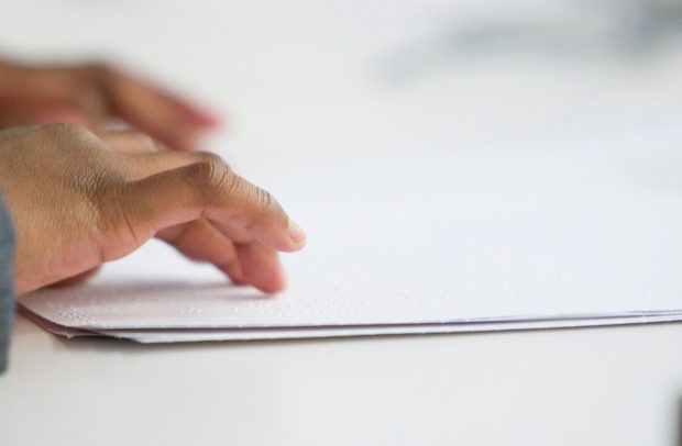 Foto parcialmente incolor da mão de uma mulher lendo uma folha com escrita em Braille. A folha e parte da mesa são brancas.