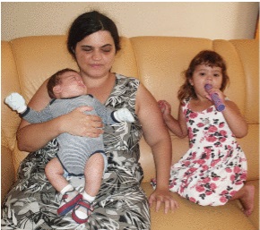 Foto de Joyce Guerra. Ela está sentada em um sofá e segura um bebê no colo. À direita dela, de joelhos no sofá, está uma menina com cerca de três anos de idade. Joyce tem pele parda, cabelo preto, longo e ondulado, que está preso. Ela usa um vestido preto com detalhes brancos.