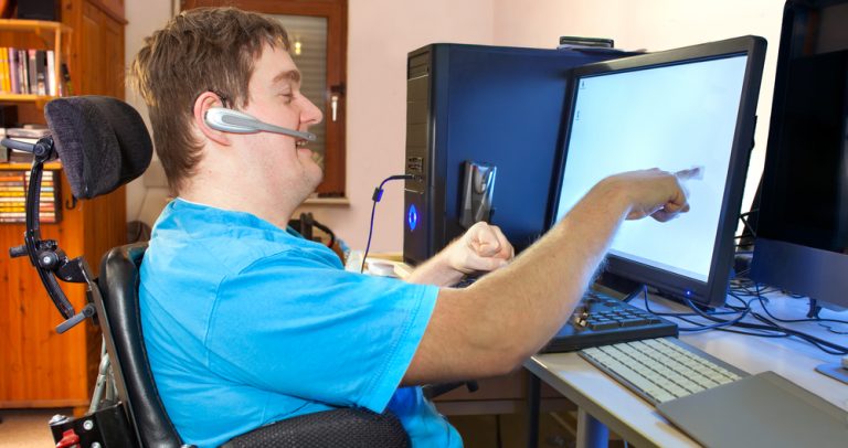 homem aponta para a tela do computador, em sua mesa de trabalho. Ele está sentado em uma cadeira de rodas e usa um recurso de tecnologia assistiva na cabeça.