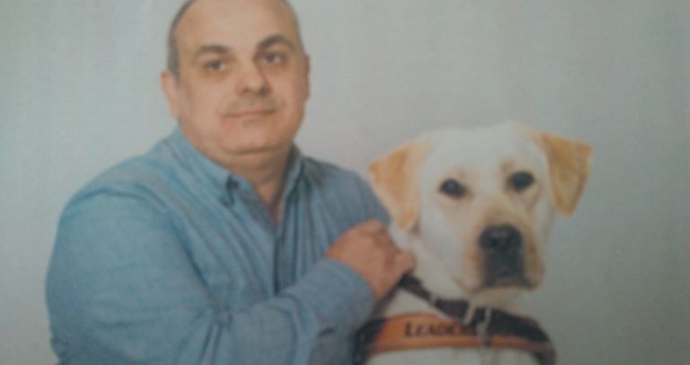 Marcelo Panico posa para foto ao lado do seu cão-guia Rudy. Marcelo tem 51 anos de idade, é branco, usa camisa de mangas compridas de cor azul. Rudy é um cão labrador branco, está com coleira marrom.