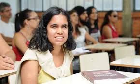Uma jovem com deficiência visual está sentada em uma carteira escolar, com materiais de estudo sobre a mesa. Ela é morena, de cabelos lisos e pretos, veste uma blusa amarela e tem um sueter branco sobre os ombros. Ela sorri. Atrás dela aparecem outros jovens sentados em suas carteiras.
