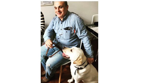 Marcelo Panico está sentado em uma cadeira, com seu cão-guia Rudy ao lado. Marcelo é branco, usa camisa e calça jeans azul claro. Rudy é um labrador branco.