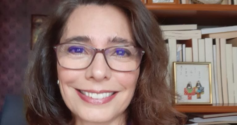 Imagem da Dra. Adriane Reis de Araújo. Ela é uma mulher branca, com cabelos escuros na altura do ombro,olhos escuros, usa óculos e está sorrindo. Está vestindo uma blusa azul sob um casaco vermelho e usa uma corrente com um pingente . Ao fundo, há uma estante de livros.