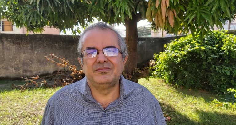 Fernando Sampaio é um homem branco, grisalho, usa óculos, e na foto está de pé, usa uma camisa azul de mangas longas dobradas, e ao fundo aparece um gramado e algumas árvores.
