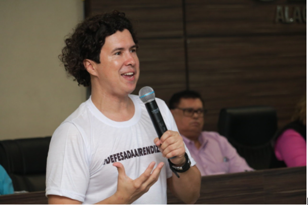 foto do auditor Leandro Carvalho. Ele está de pé falando ao microfone, em uma sala de auditório. É um homem branco, com cabelos encaracolados de corte abaixo da orelha, usa uma camiseta branca com as palavras em letras pretas: Defesa da aprendizagem