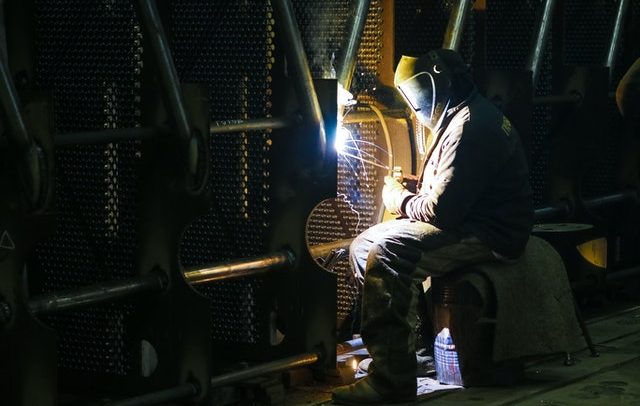 Homem em ambiente de indústria metalúrgica utiliza uniforme e capacetr. Ele está sentado e aponta um foco de luz para o local onde está realizando uma solda
