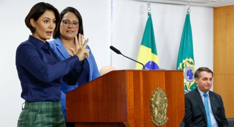 Primeira-dama Michelle Bolsonaro faz discurso em LIBRAS, em um púlpito de madeira com o brasão do governo federal. Michelle é uma mulher branca, de cabelos curtos pretos, olhos escuros, veste uma camisa azul e uma saia xadrez verde. Ao seu lado está uma mulher branca, cabelos pretos curtos e veste um casaco azul. Ao lado, está sentado em uma cadeira o presidente Jair Bolsonaro. Atrás, é possível ver as bandeiras do Brasil e da independência