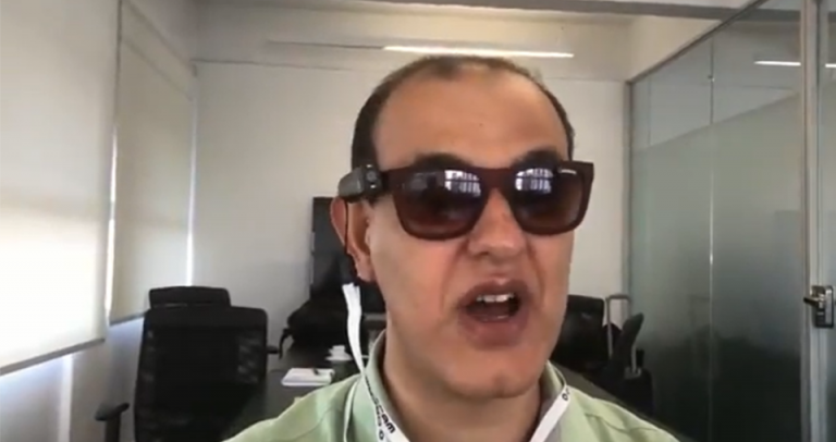 Beto Pereira é um homem calvo, usa óculos escuros. Ele está vestindo uma camisa social clara, da qual aparece só a gola.