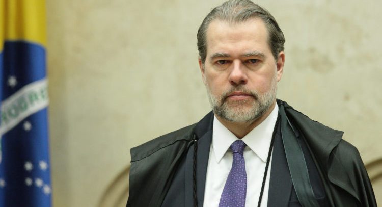 Foto do Ministro Dias Toffoli, um homem com cabelo, barba e bigode grisalhos, olhos castanhos. Ele está vestindo uma camisa social branca, terno e gravata azul-marinho, e, por cima, uma capa preta, própria para os juízes. Ao fundo, é possível ver um pedaço da bandeira do Brasil.