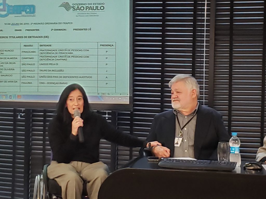 A imagem mostra Chico Pirata com com a Secretária Estadual dos Direitos da Pessoa com Deficiência, Célia Leão. Chico é um homem branco, grisalho, de barba, está usando um tesno escuro com camisa cinza, sentado atrás de uma mesa com teclado e uma garrafa de água. Ao seu lado está Célia Leão, uma mulher branca, de cabelos e olhos escuros, veste uma blusa preta e calça marrom. Ela é cadeirante e está segurando um microfone.