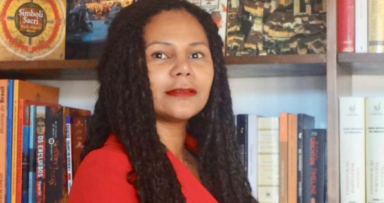 Valdirene Assis é uma mulher morena, de cabelos longos, escuros e cacheados. Os olhos escuros e usa batom vermelho. Veste um blazer vermelho. Atrás dela, há uma estante com muitos livros.