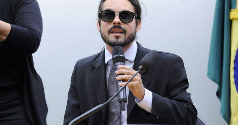 Rafael Giguer é um homem branco, de barba e cabelos pretos, usa óculos escuros. Está vestindo um terno escuro com camisa azul clara e gravata mais escura. Está sentado em uma mesa com uma placa onde está escrito seu nome e cargo.
