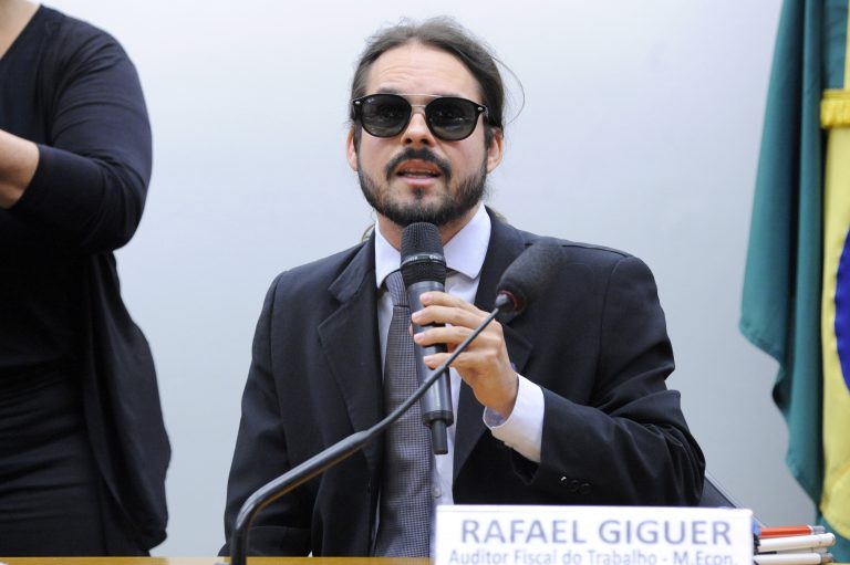 Rafael Giguer é um homem branco, de barba e cabelos pretos, usa óculos escuros. Está vestindo um terno escuro com camisa azul clara e gravata mais escura. Está sentado em uma mesa com uma placa onde está escrito seu nome e cargo.