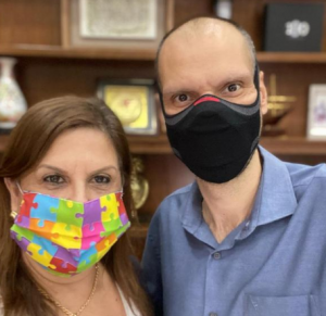 Foto de Sílvia Grecco com o prefeito Bruno Covas. Do lado direito está Silvia, que tem cabelos castanhos claros e olhos castanhos. Ela usa uma máscara colorida com desenhos de paças de quebra-cabeças. Do lado direito está Bruno Covas, um homem calvo, de olhos escuros. Ele está usando uma camisa social azul clara e uma máscara preta com detalhes em cinza e vermelho.
