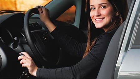 Uma mulher branca, de cabelos pretos e longos, está sentada e segurando ao volante e na alavanca de freio de um carro adaptado. Ela está sorrindo.