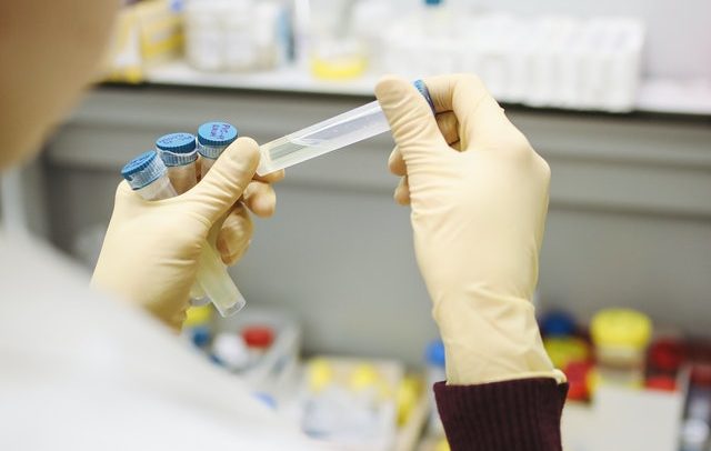 Imagem focaliza nas mãos de uma pessoa em um laboratório. Ela veste luvas de cor bege e segura frascos transparentes, como amostras de vacina.