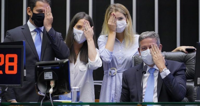 quatro pessoas, dois homens e duas mulheres, uma do lado da outra. Somente o último está sentado. O primeiro homem é moreno, de cabelos curtos pretos, veste terno preto, com camisa branca, gravata azul e usa máscara preta. Ao seu lado está uma mulher branca, de cabelos longos pretos, usando uma camisa e uma máscara branca. Logo após, uma mulher loira, de vestido azul listrado, usa uma máscara branca. Por último (sentado, está um homem grisalho, branco, de terno escuro, camisa branca e gravata azul. Todos as pessoas na foto estão cobrindo um dos olhos com a mão, sinalizando a visão monocular.