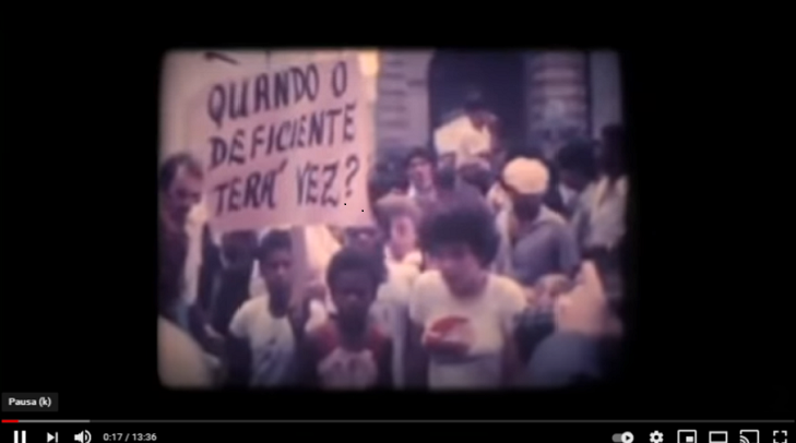 Reprodução de imagem do documentário no Youtube, que mostra uma foto antiga com um manifestantes reunidos portando uma placa onde está escrita a frase: quando o deficiente terá vez?