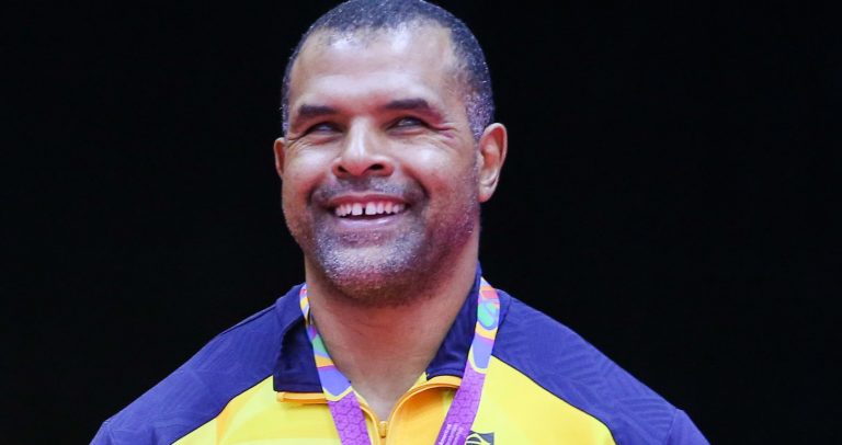 Antonio Tenório é um homem negro, de cabelos curtos e grisalhos. Ele usa uma camisa amarela com uma faixa azul nos ombros e gola azul. No peito, tem uma pequena imagem da bandeira do Brasil. Ele está sorrindo.