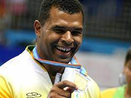 Foto do judoca Antônio Tenório. Ele é um homem negro, com cabelos pretos bem curtos, olhos escuros. Está sorrindo e mostrando sua medalha de ouro, que está pendurada em seu pescoço.