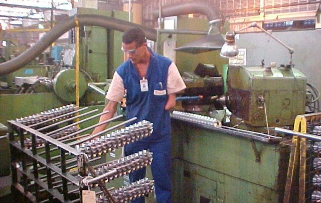 Imagem de um trabalhador com deficiência em uma indústria metalúrgica. Ele é um homem negro, de cabelos escuros e curtos, utiliza um óculos de proteção. Veste um macacão azul com mangas curtas em tom de bege, e usa um crachá no pescoço. Ele possui o braço esquerdo encurtado e sem mão. Com o braço direito, ele manuseia uma máquina.