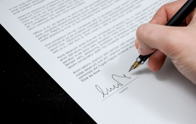 A foto detalha a mão de uma pessoa assinando um documento com uma caneta de cor preta.