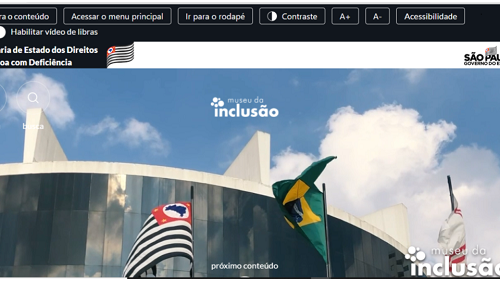 Reprodução da página principal do site do Museu da Inclusão. Na barra superior, há botões “ir para o conteúdo”, “acessar o menu principal”, ir para o rodapé”, “contraste A+ A-, “Acessibiildade. Abaixo, há o botão escrito “habilitar vídeo de libras” e o título Secretaria do Estado dos Direitos da pessoa com deficiência, junto à bandeira de São Paulo. Abaixo, há uma foto detalhando um céu azul e nuvens brancas, e um pedaço da parte superior da fachada do Memorial da América Latina, onde se localiza o Museu da Inclusão. Há 3 bandeiras hasteadas. No canto direito, há o logo do memorial da Inclusão