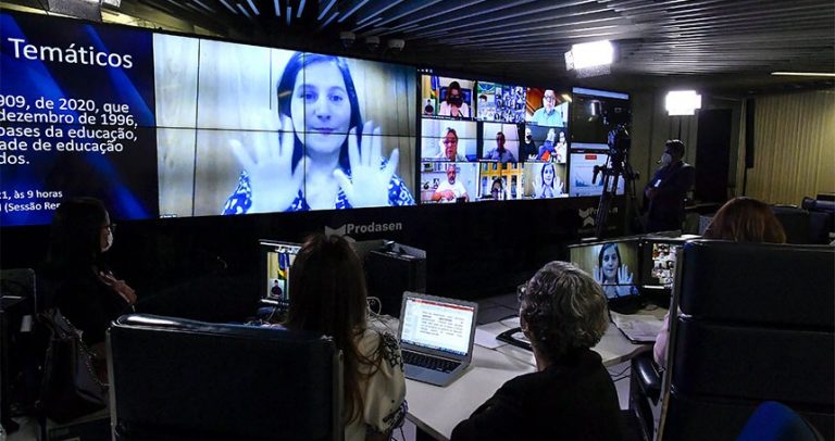 Imagem mostra sala escura com telão que mostra a intérprete de libras em um quadro e a imagem dos participantes no outro. À frente dele, estão sentadas três pessoas de costas, voltadas para o telão, em uma mesa com notebooks.